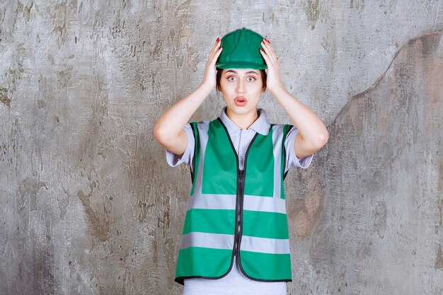 Vrouwelijke ingenieur in groen uniform en helm die hoofd vasthoudt en ziet er doodsbang uit