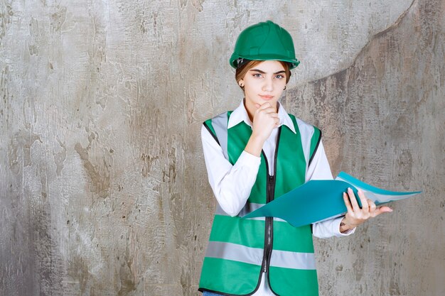 Vrouwelijke ingenieur in groen uniform en helm met een groene projectmap, denken en analyseren.