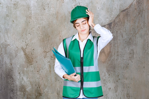 Vrouwelijke ingenieur in groen uniform en helm met een groene projectmap en ziet er moe en slaperig uit