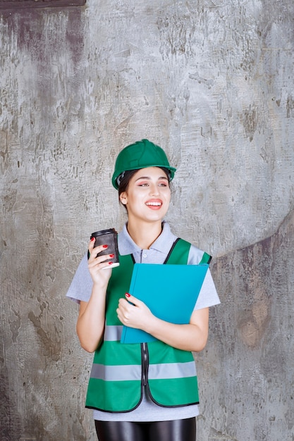 Vrouwelijke ingenieur in groen uniform en helm met een zwarte koffiekopje en een blauwe projectmap.