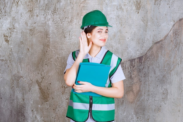 Vrouwelijke ingenieur in groene helm die een blauwe map vasthoudt en oor opent om goed te horen