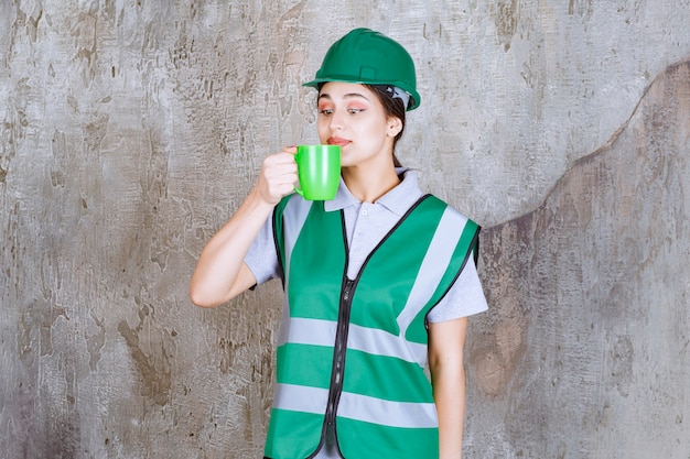 Vrouwelijke ingenieur in groene helm die een groene koffiemok houdt en het product ruikt.