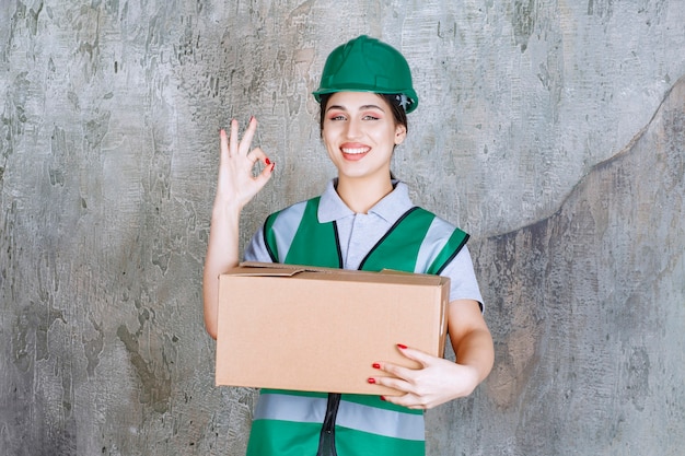 Vrouwelijke ingenieur in groene helm die een kartonnen doos vasthoudt en een tevredenheidsteken toont