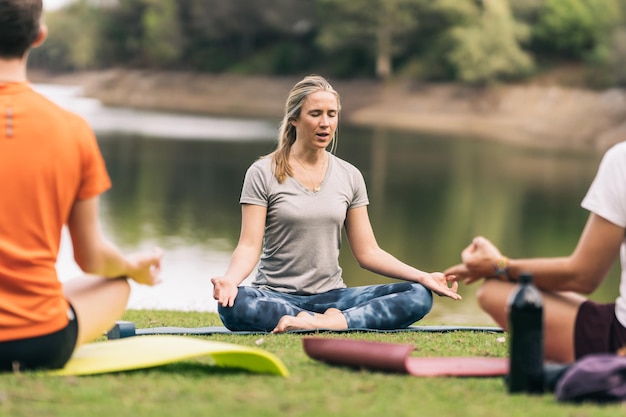 Vrouwelijke instructeur zittend op een mat die de lotushouding doet tijdens een yogales in een buitenpark