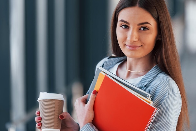 Vrouwelijke jonge student is in de gang van een universiteit met notitieblokken en een kopje drank