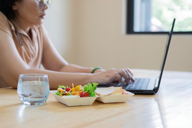 Vrouwelijke kantoormedewerker zit aan een bureau met een laptop en een glas water met gezond voedsel in een doos