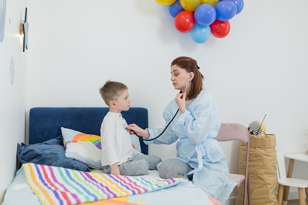 Vrouwelijke kinderarts onderzoekt een zieke kleine jongen bij hem thuis, in een poging de patiënt te vermaken en op te vrolijken