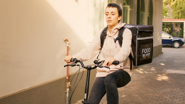 Vrouwelijke koerier die naar de voordeur van de klant gaat en lunchmaaltijd levert, fietst om klanten afhaalmaaltijden te geven in thermische rugzak. Vrouw aan het werk als vervoerder op de fiets, zonnige dag.