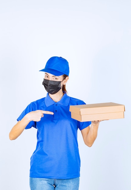 Vrouwelijke koerier in blauw uniform en gezichtsmasker die een kartonnen doos aan de klant levert.