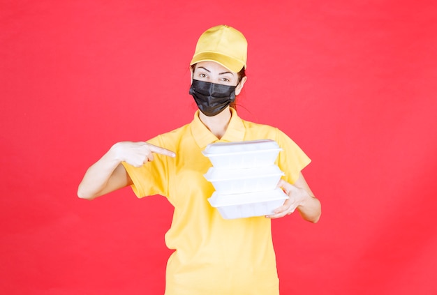 Vrouwelijke koerier in geel uniform en zwart masker die meerdere afhaalpakketten vasthoudt en de handtekeninglijst presenteert