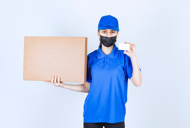 Vrouwelijke koerier in masker en blauw uniform die een groot kartonnen pakket houdt en haar visitekaartje voorstelt.