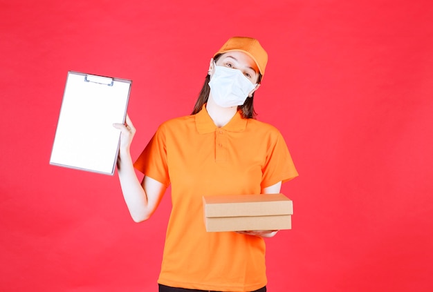 Vrouwelijke koerier in oranje kleur uniform en masker met een kartonnen doos en presenteert de lijst ter ondertekening.