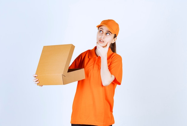 Vrouwelijke koerier in oranje uniform met een open kartonnen doos en ziet er verward en attent uit