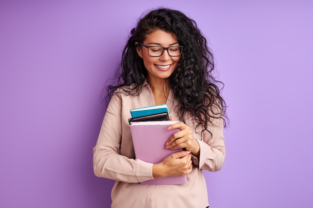 Vrouwelijke leraar met boeken in handen geïsoleerd op paarse muur, jonge dame in bril geniet van onderwijs, lach