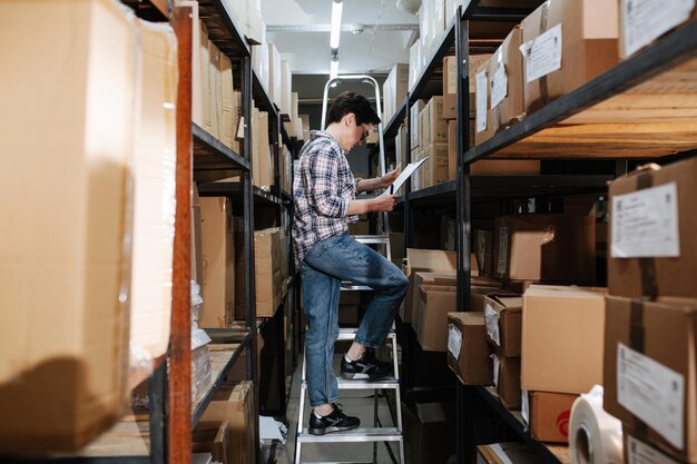 Vrouwelijke magazijnmedewerker lezen die op de ladder naast planken staat