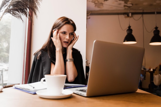 Vrouwelijke manager met sterke hoofdpijn die op afstand online aan laptop werkt in loftcafé Droevig meisje met laptop kreeg migraine raakt haar hoofd aan vanwege pijn