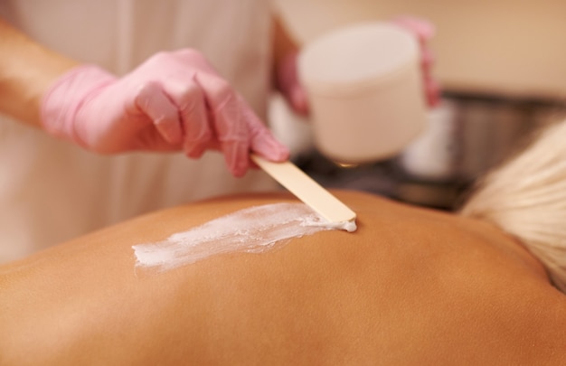 Vrouwelijke masseur past een massagecrème scrub toe met behulp van een houten spatel op de rug van een jonge vrouw die op een massagetafel ligt en geniet van een therapiesessie in de spa-salon