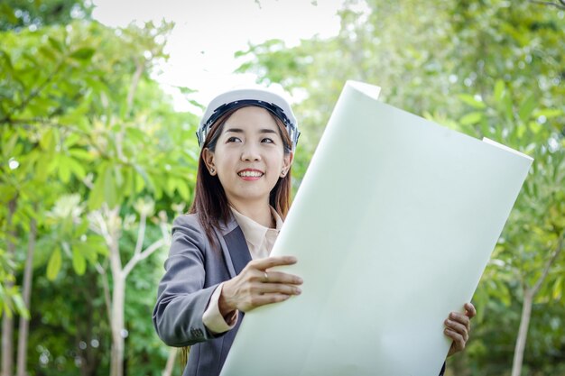 Vrouwelijke milieu-ingenieur draagt een pak, draagt een witte hoed, staande houden een papieren plan