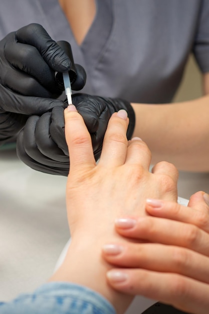 Vrouwelijke nagels lakken. Handen van manicure in zwarte handschoenen past transparante nagellak toe op vrouwelijke nagels in een manicuresalon.