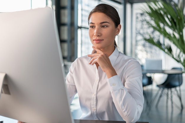 Vrouwelijke officemanager in wit overhemd op kantoor interieur achtergrond mooie vrouw kantoormedewerker vrouw die werkt met computer in groot kantoor concept van werk