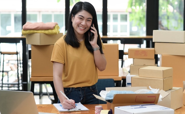 Vrouwelijke online verkoop belt naar de telefoon en maakt aantekeningen en neemt bestellingen op met een glimlach, de pakketdoos wordt op tafel geplaatst, ze kijkt naar de camera en glimlacht.