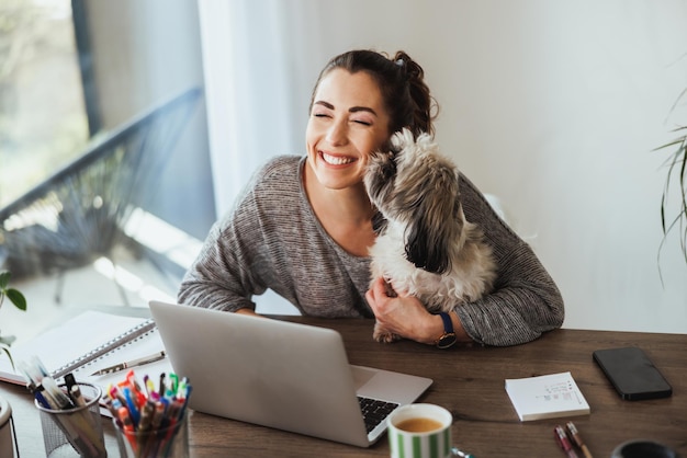 Vrouwelijke ontwerper die plezier heeft met haar hond terwijl ze aan een laptop werkt in haar kantoor aan huis.