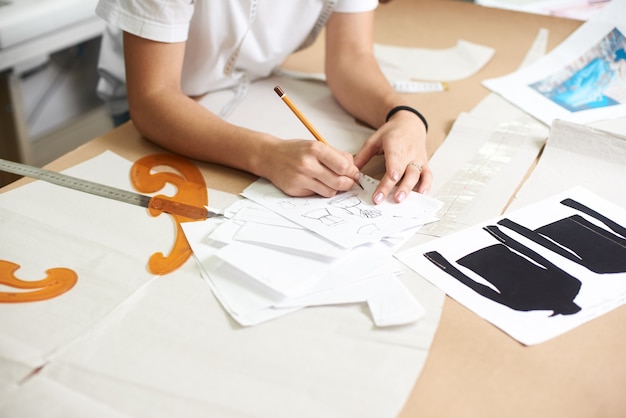 Foto vrouwelijke ontwerper tekening potloodschetsen van kleding zittend aan grote tafel met platte papieren patronen