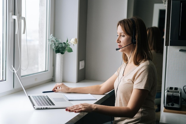 Vrouwelijke operator praten met behulp van headset en raadpleging van klant zittend op de vensterbank met laptop vanuit kantoor aan huis