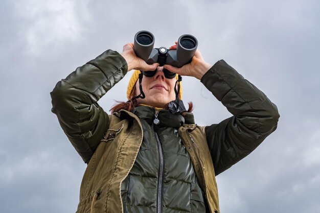 Vrouwelijke ornitholoog birdman kijkt naar vogels met een verrekijker op een vuil grindweg tegen de achtergrond van een stormachtige lucht