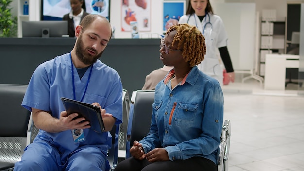 Vrouwelijke patiënt en medisch assistent die naar een digitale tablet kijkt om te praten over de diagnose van de gezondheidszorg in de wachtruimte bij de receptie van het ziekenhuis. Diverse mensen doen controle examen in wachtkamer.