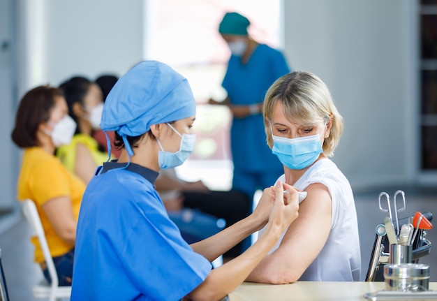 Foto vrouwelijke professionele arts draagt een gezichtsmasker en een blauw ziekenhuisuniform dat een coronovirusvaccin-spuitnaald injecteert in een blanke senior vrouwelijke patiëntschouder aan het bureau van de kliniek.