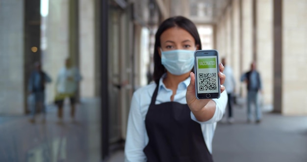 Vrouwelijke restauranteigenaar met veiligheidsmasker die mobiele telefoon met gevaccineerde qrcode naar de camera laat zien