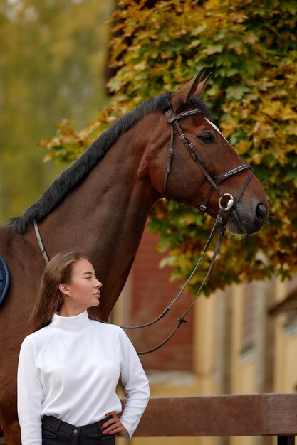 Vrouwelijke ruiter die met haar bruin volbloed paard in stal gaat Concept van dierenverzorging Landelijke rust en vrije tijd Idee van groen toerisme Jonge Europese vrouw die helm en uniform draagt