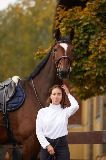 Vrouwelijke ruiter die met haar bruin volbloed paard in stal gaat Concept van dierenverzorging Landelijke rust en vrije tijd Idee van groen toerisme Jonge Europese vrouw die helm en uniform draagt