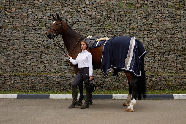 Vrouwelijke ruiter die met haar bruin volbloed paard in stal gaat Concept van dierenverzorging Landelijke rust en vrije tijd Idee van groen toerisme Jonge Europese vrouw die helm en uniform draagt