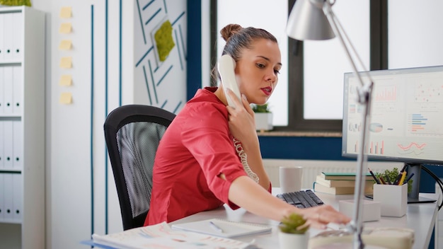 Vrouwelijke secretaresse die vaste telefoon gebruikt om te bellen van manager, bezig met bedrijfsontwikkeling met financiële grafieken. Projectstrategie voor kantoormedewerkers met verkoopstatistieken en onderzoek.