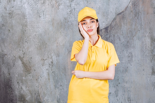 Vrouwelijke serviceagent in geel uniform staande op betonnen muur en ziet er verward en attent uit.