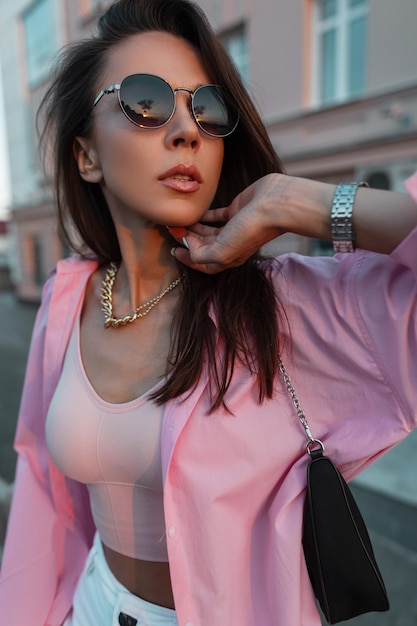 Vrouwelijke stijlvolle portret van een mooie jonge glamoureuze meisje met ronde vintage zonnebril in een modieus roze shirt en top met een portemonnee en een gouden ketting wandelen en poses in de stad bij zonsondergang