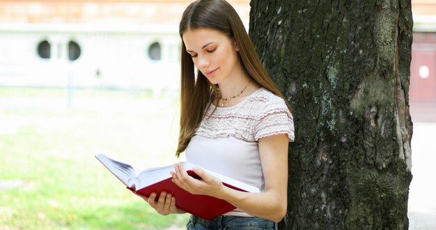 Vrouwelijke student die een boek openlucht in het park lezen die tegen een boom en het glimlachen liggen