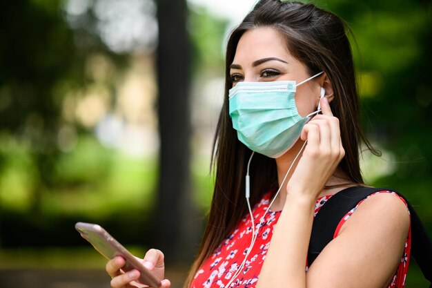 Vrouwelijke student loopt in een park terwijl ze haar smartphone gebruikt om naar muziek te luisteren en een masker draagt om zich te beschermen tegen de coronavirus covid-19 pandemie