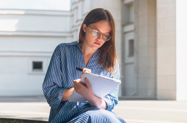 Vrouwelijke student portret zitten in de buurt van de universiteit en leren schrijven smth lezen met Kladblok op...