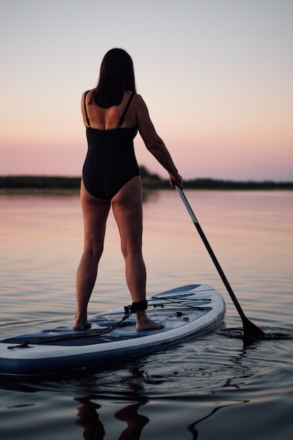 Vrouwelijke sup boarder van middelbare leeftijd stuurbord met peddel vooruitkijkend op meer met verbazingwekkende roze lucht op de achtergrond in zwembroek in de zomer Actieve levensstijl