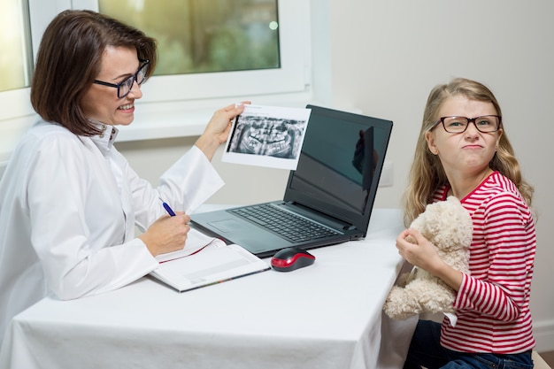 Vrouwelijke tandartsorthodontist met röntgenfoto die aan een kind spreekt.
