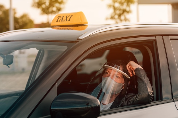 Vrouwelijke taxichauffeur met beschermend gezichtsmasker en plastic vizier wachtend in de auto