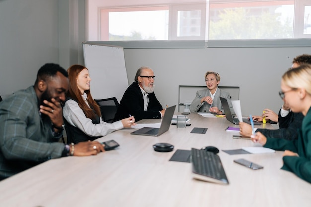 Foto vrouwelijke teamleider van middelbare leeftijd zit aan tafel met multiraciale collega's en bespreekt werkvraagstukken tijdens onderhandelingen.