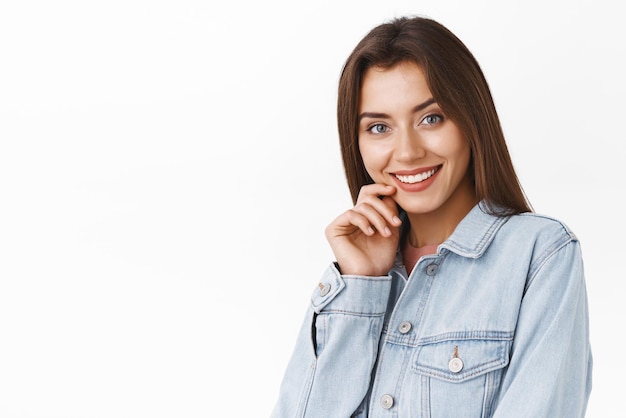 Vrouwelijke tedere en mooie brunette vrouw in spijkerjasje aanraken lip dwaze en sensuele glimlach stralend van vreugde en liefde staande witte achtergrond verrukt blij met leuk gesprek