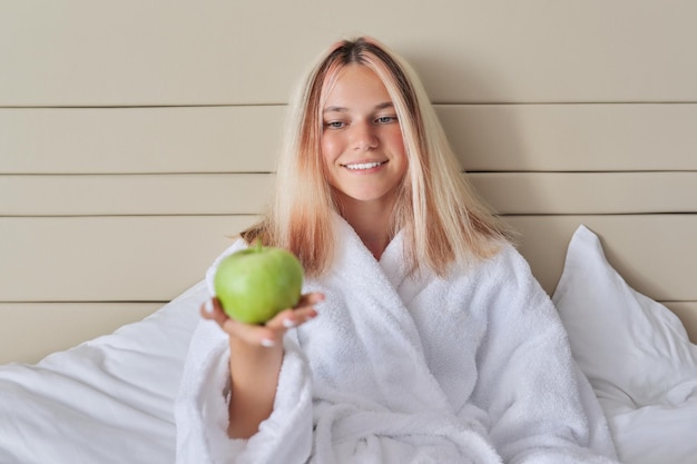 Vrouwelijke tiener met groene appel in witte badjas zittend in bed