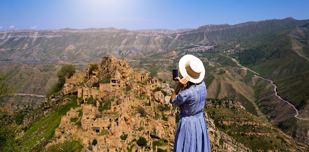 Vrouwelijke toerist hoog in de bergen van Dagestan bewondert het adembenemende panoramische uitzicht op de natuur en de oude verlaten stad Gamsutl. Maakt foto's van attracties en ruïnes, selfies op een smartphone