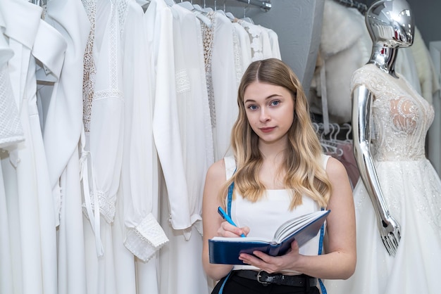 Vrouwelijke verkoopster die een notitieboekje vasthoudt en poseert in een salon die trouwjurken verkoopt