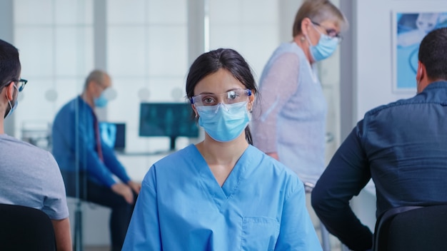 Vrouwelijke verpleegster met gezichtsmasker tegen coronavirus in de wachtruimte van het ziekenhuis die naar de camera kijkt. Senior vrouw met een handicap lopen met behulp van zimmer frame.